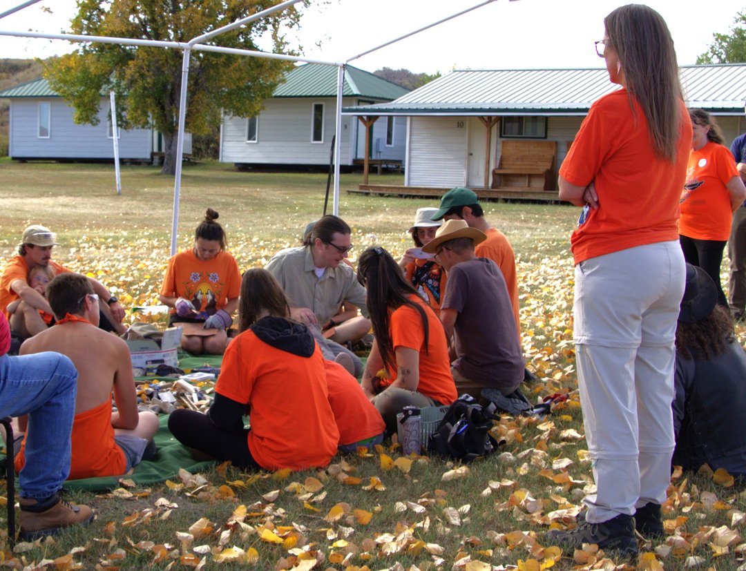 A group of people sits in a circle under a canopy frame on a grassy area covered with fallen leaves. Most wear orange shirts, and white cabins with green roofs are visible in the background along with a tree with autumn foliage.