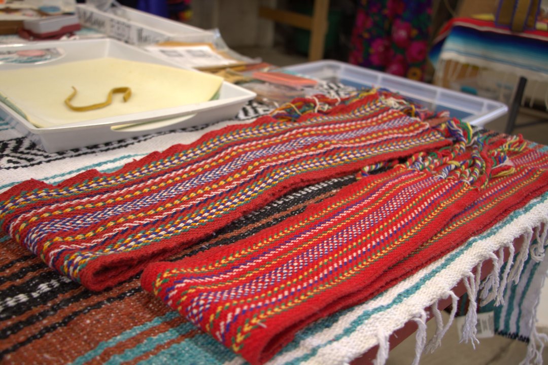 Two L'Assomption sash with vibrant red, yellow, blue, and white patterns are laid out on a table covered with a striped textile.