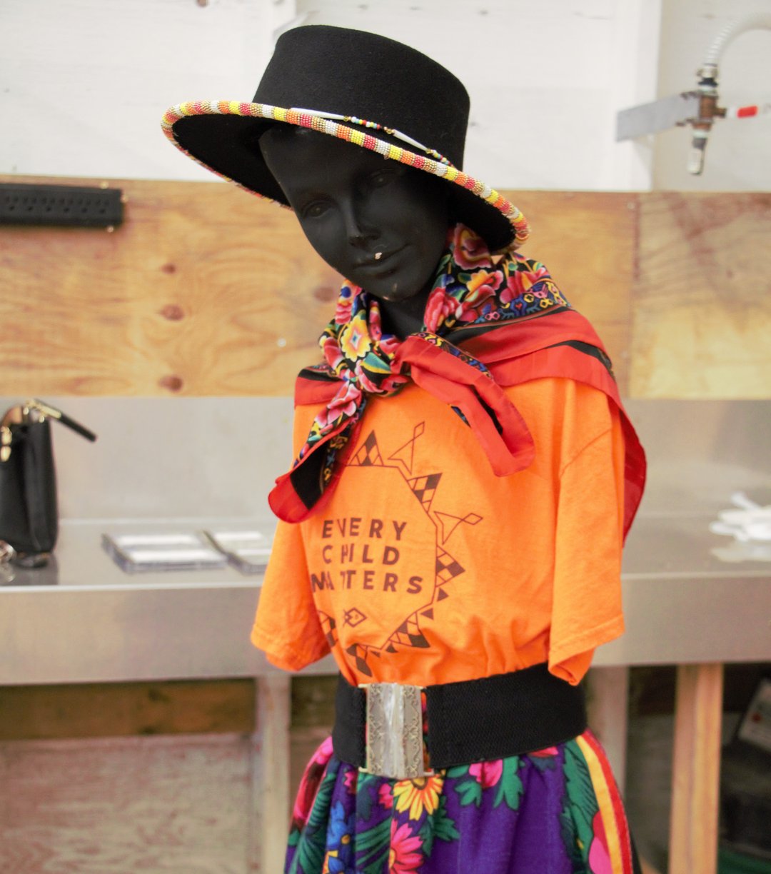 A mannequin dressed in an orange “Every Child Matters” T-shirt, a colorful floral skirt, and a wide black hat with a matching patterned scarf.