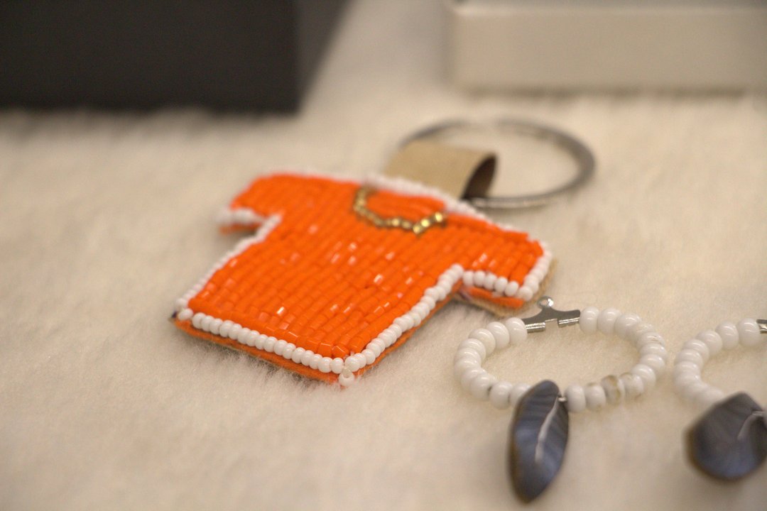 A small beaded keychain shaped like an orange shirt with white bead trim and a gold necklace design, symbolizing “Every Child Matters.” Next to it are two circular beaded pieces with feather-shaped pendants, placed on a soft white surface.