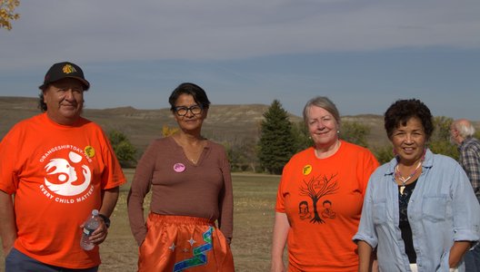 Four people standing outdoors on grass with trees and hills in the background. Two wear bright orange shirts with designs and text, one reading “Every Child Matters,” while another wears a orange ribbon skirt and one a light blue shirt.