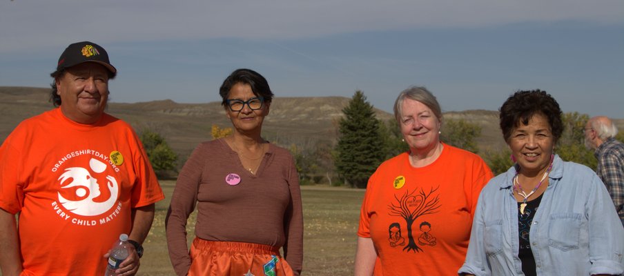 Four people standing outdoors on grass with trees and hills in the background. Two wear bright orange shirts with designs and text, one reading “Every Child Matters,” while another wears a orange ribbon skirt and one a light blue shirt.