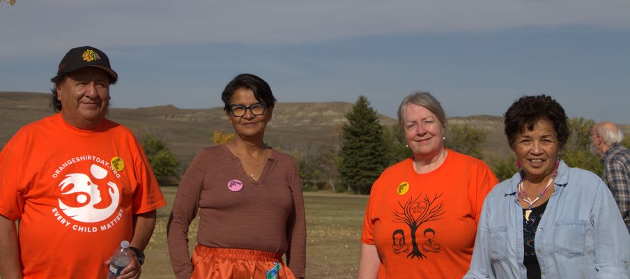 Four people standing outdoors on grass with trees and hills in the background. Two wear bright orange shirts with designs and text, one reading “Every Child Matters,” while another wears a orange ribbon skirt and one a light blue shirt.