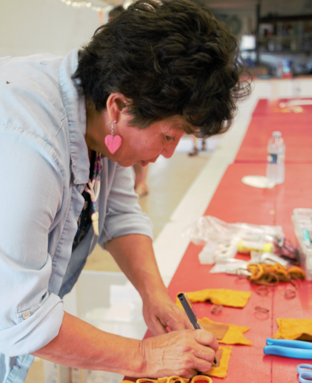 Person at a table working on a craft project with leather pieces and string. The table has various supplies including scissors, pliers, plastic bags, and other crafting materials arranged on a red surface.