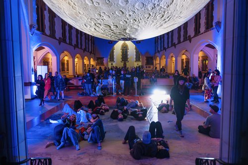 A photo of people sitting down looking at a big art installation of a moon at Nuit Blanche YXE 2023.