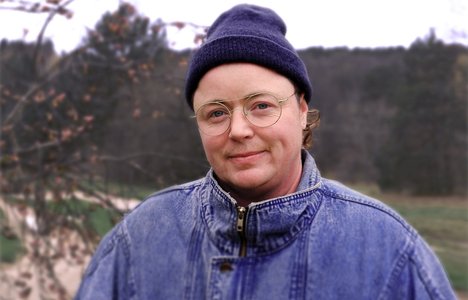 Portrait of artist Lou Sheppard standing outdoors in front of a softly blurred landscape. Lou wears round glasses, a dark knit cap, and a blue denim jacket, with bare tree branches and a wooded background visible behind him.