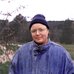 Portrait of artist Lou Sheppard standing outdoors in front of a softly blurred landscape. Lou wears round glasses, a dark knit cap, and a blue denim jacket, with bare tree branches and a wooded background visible behind him.