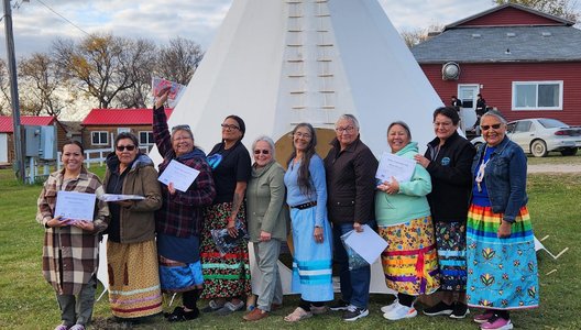 A group of nine people standing in front of a large white tipi on a grassy area. They are holding papers and wearing colorful ribbon skirts and varied clothing.