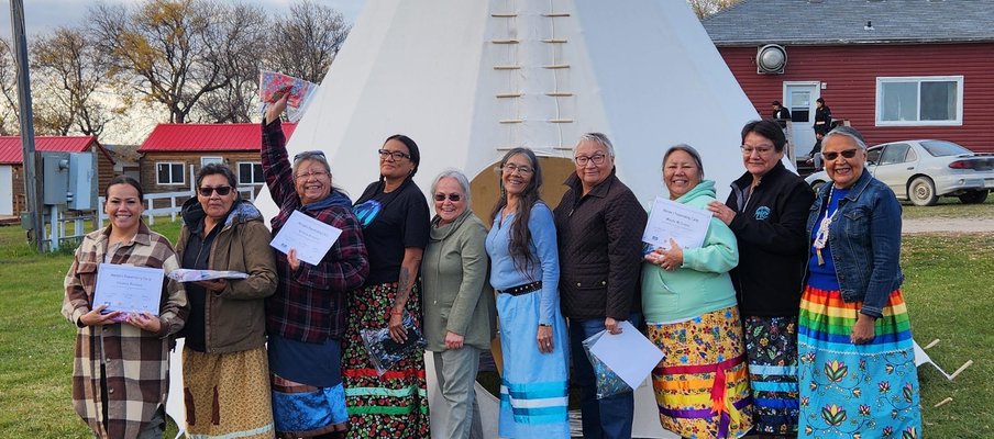 A group of nine people standing in front of a large white tipi on a grassy area. They are holding papers and wearing colorful ribbon skirts and varied clothing.
