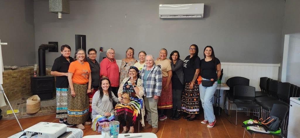 Thirteen people standing indoors in a semi-circle with one person seated in front. Several wear colorful ribbon skirts, and the room has gray walls, a wood stove, chairs, and a table with a projector in the foreground.
