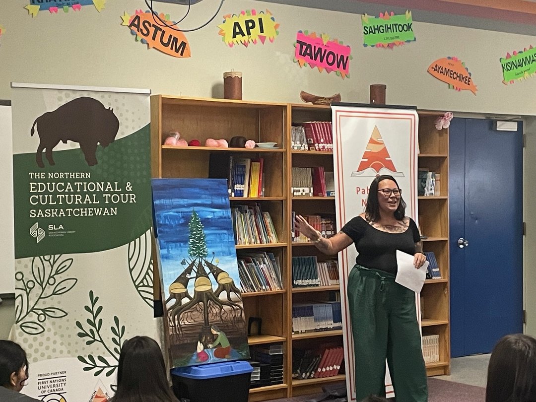 A person stands in front of bookshelves, speaking and gesturing toward a painting of tipis and a tree.