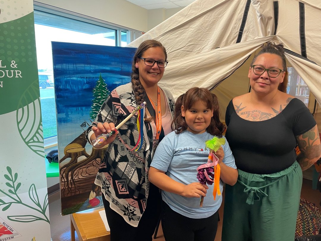 Three people stand indoors in front of a large canvas painting of tipis and a tree, next to a beige canvas tent. One person holds a decorated craft, and the child in the center holds colorful craft items with feathers and ribbons.