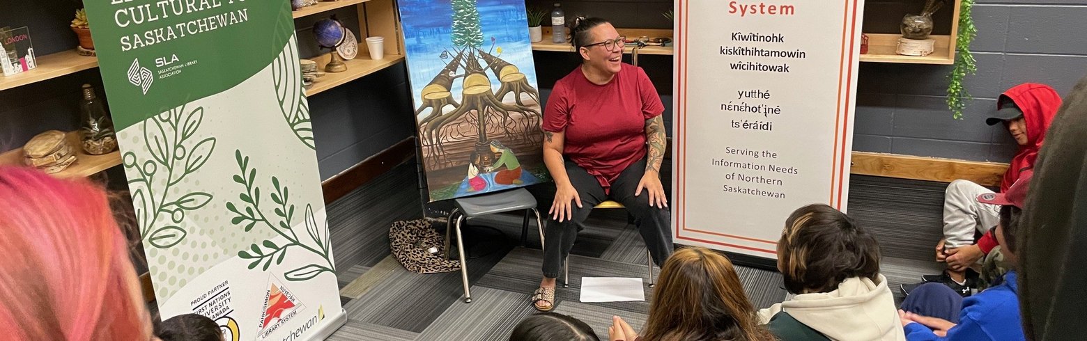 A person is seated in a library space speaking to a group of children sitting on the floor. Behind them are two banners about the Northern Educational & Cultural Tour and the Pahkisimon Nuyeáh Library System, along with a painting of tipis and a tree.