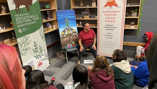 A person is seated in a library space speaking to a group of children sitting on the floor. Behind them are two banners about the Northern Educational & Cultural Tour and the Pahkisimon Nuyeáh Library System, along with a painting of tipis and a tree.