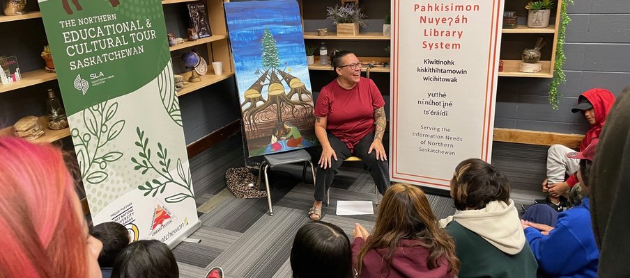 A person is seated in a library space speaking to a group of children sitting on the floor. Behind them are two banners about the Northern Educational & Cultural Tour and the Pahkisimon Nuyeáh Library System, along with a painting of tipis and a tree.