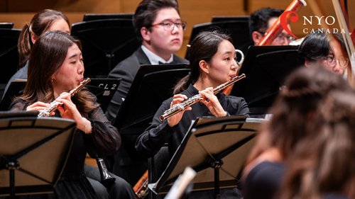 Two flutists in focus, playing their instruments onstage, surrounded by the rest of the woodwind section in performance.