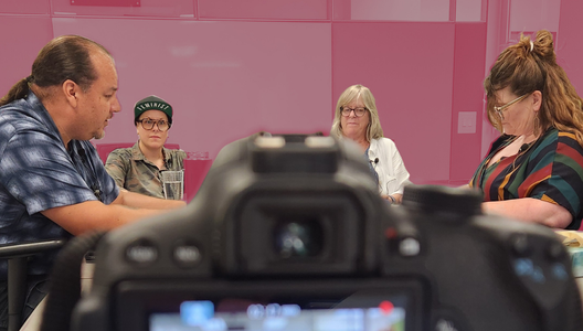 Photo of a camera taking a photo of four people who are doing a podcast in a office.