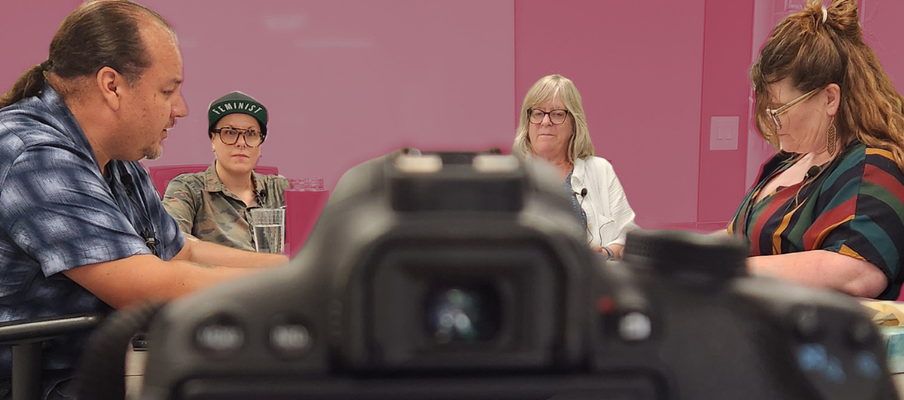 Photo of a camera taking a photo of four people who are doing a podcast in a office.