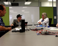 Four people sitting around a conference table with papers, water glasses, a magazine, and audio recording equipment. A whiteboard and green wall panels are in the background.