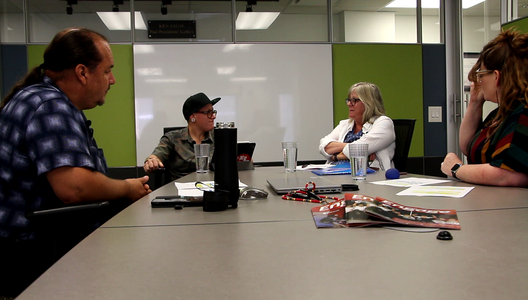 Four people sitting around a conference table with papers, water glasses, a magazine, and audio recording equipment. A whiteboard and green wall panels are in the background.