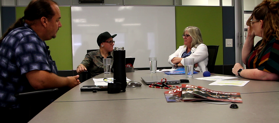 Four people sitting around a conference table with papers, water glasses, a magazine, and audio recording equipment. A whiteboard and green wall panels are in the background.