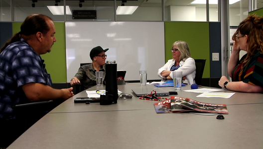 Four people sitting around a conference table with papers, water glasses, a magazine, and audio recording equipment. A whiteboard and green wall panels are in the background.