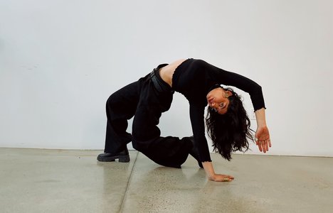 Rawda Mabrouk dressed in black strikes a dramatic, arched pose on a polished concrete floor against a plain white wall. One hand and one knee support the body while the torso curves backward, creating a sculptural, dance-like shape.