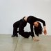 Rawda Mabrouk dressed in black strikes a dramatic, arched pose on a polished concrete floor against a plain white wall. One hand and one knee support the body while the torso curves backward, creating a sculptural, dance-like shape.
