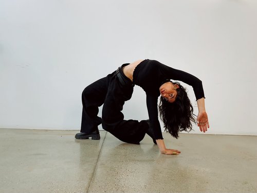 Rawda Mabrouk dressed in black strikes a dramatic, arched pose on a polished concrete floor against a plain white wall. One hand and one knee support the body while the torso curves backward, creating a sculptural, dance-like shape.