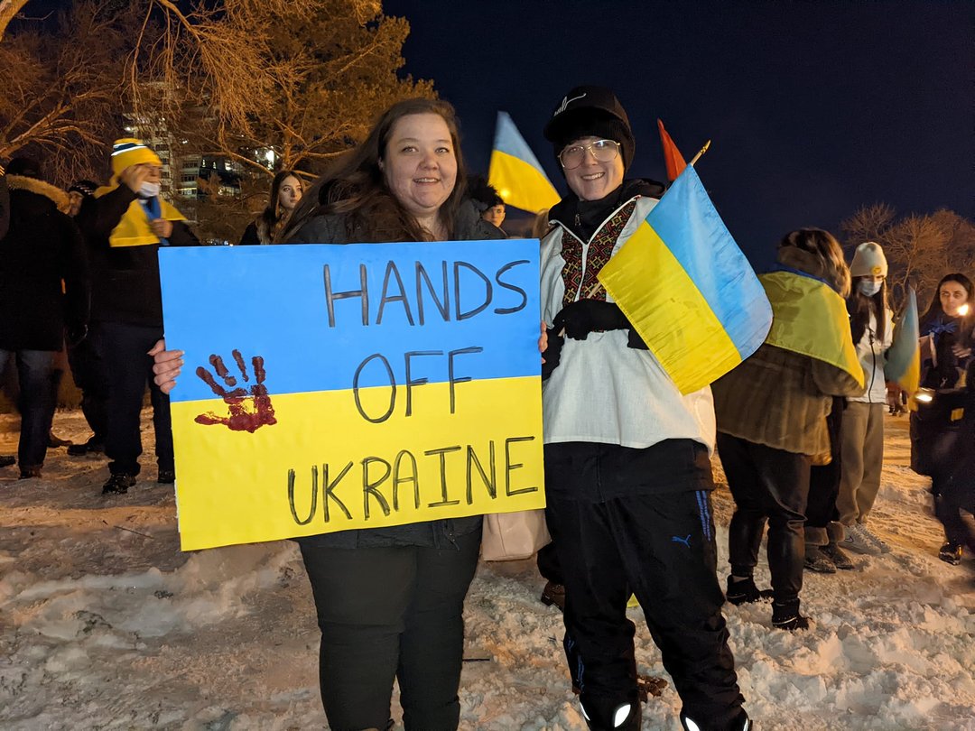 Two people stand outdoors at night on snow-covered ground during a rally, holding a large blue and yellow sign that reads “HANDS OFF UKRAINE.” One person is also holding two Ukrainian flags, and several other participants with flags are visible in the background.