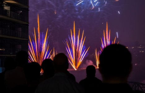 Silhouetted spectators watch a nighttime fireworks display, with bright blue and orange bursts lighting the sky. The colorful pyrotechnics rise above the water beside nearby buildings, creating a dramatic urban scene.