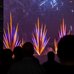 Silhouetted spectators watch a nighttime fireworks display, with bright blue and orange bursts lighting the sky. The colorful pyrotechnics rise above the water beside nearby buildings, creating a dramatic urban scene.