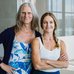 Miki Mappin and Kyle Syverson stand side by side in a bright studio space, facing the camera. Large windows and soft natural light frame the portrait, with one person wearing a floral-patterned outfit and the other dressed in a light tank top and dark pan