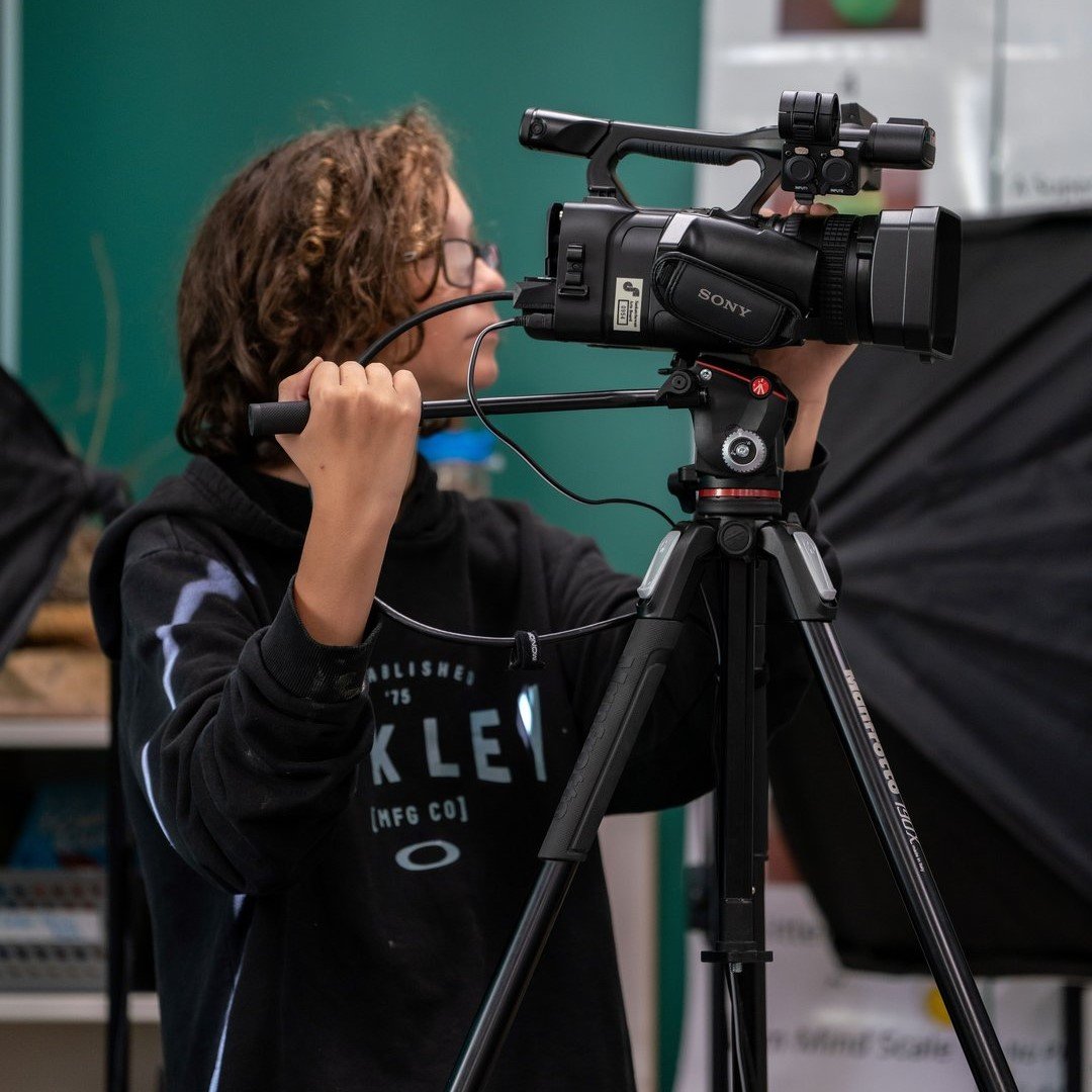 Person standing indoors operating a professional video camera mounted on a tripod.