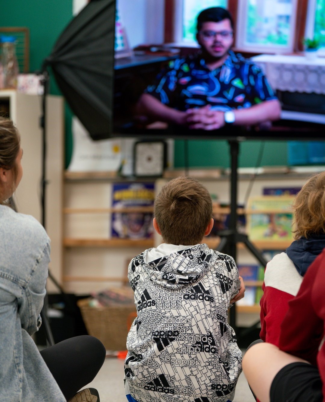 Children sitting on the floor in a classroom, watching a large monitor that displays a person speaking.