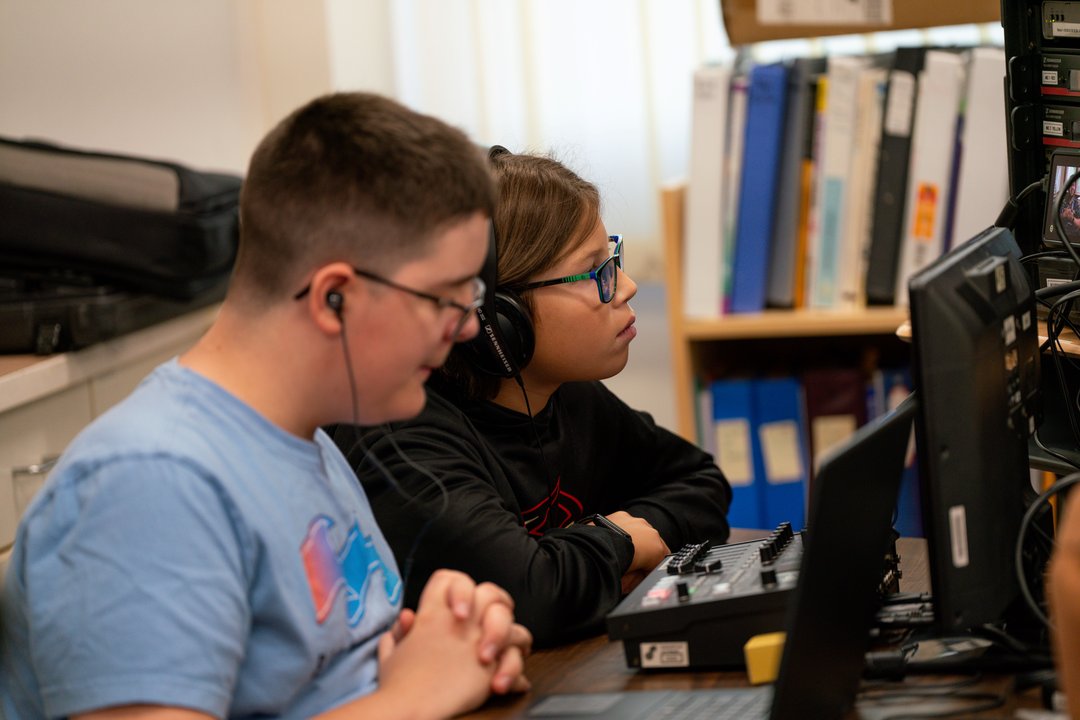 Two people sitting at a table operating audio or video equipment. One person is wearing headphones and adjusting controls on a sound mixer, while the other is seated nearby with hands clasped.