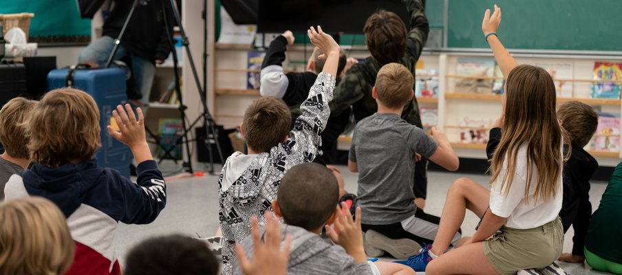 A group of children sitting on the floor in a classroom, facing a large monitor and raising their hands. Bookshelves line the back wall, and a person with a camera and lighting equipment is visible in the corner.
