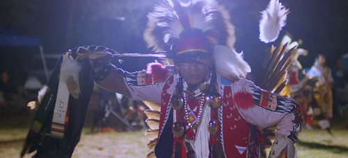 Still shot from the film with a Indigenous man dressed in traditional Indigenous dress dancing at a Powwoww..