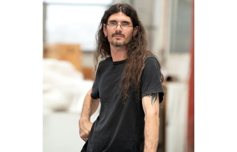 Portrait of the artist, d harding,with long dark hair and glasses, wearing a black T-shirt and standing indoors. The person faces slightly to the side with one hand at their hip, while a softly blurred studio or workshop setting appears in the background.