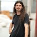 Portrait of the artist, d harding,with long dark hair and glasses, wearing a black T-shirt and standing indoors. The person faces slightly to the side with one hand at their hip, while a softly blurred studio or workshop setting appears in the background.