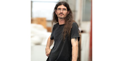 Portrait of the artist, d harding,with long dark hair and glasses, wearing a black T-shirt and standing indoors. The person faces slightly to the side with one hand at their hip, while a softly blurred studio or workshop setting appears in the background.