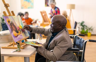 A Black man who is using a wheelchair painting a colourful still-life in a group class setting.