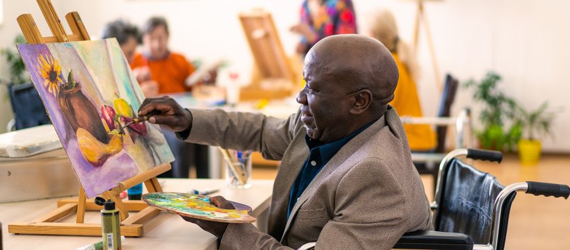 A Black man who is using a wheelchair painting a colourful still-life in a group class setting.
