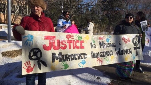 A group of people stand outdoors on a snowy sidewalk holding a large banner that reads “Justice for Missing & Murdered Indigenous Women.” The banner is decorated with colorful handprints and symbols, and some participants hold additional signs in the back