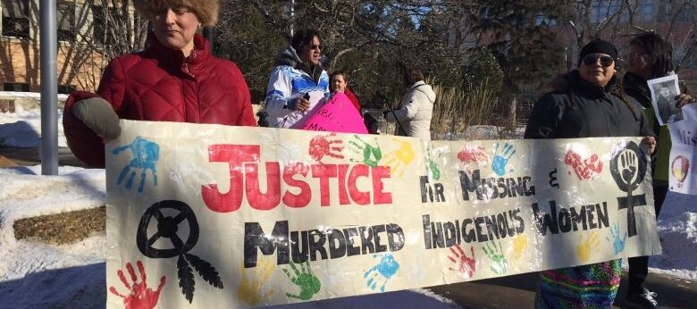 A group of people stand outdoors on a snowy sidewalk holding a large banner that reads “Justice for Missing & Murdered Indigenous Women.” The banner is decorated with colorful handprints and symbols, and some participants hold additional signs in the back