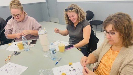 Three people seated at a table working on a craft activity with string and small materials. The table has printed instructions, water bottles, cups, and a phone.