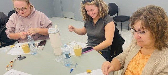 Three people seated at a table working on a craft activity with string and small materials. The table has printed instructions, water bottles, cups, and a phone.